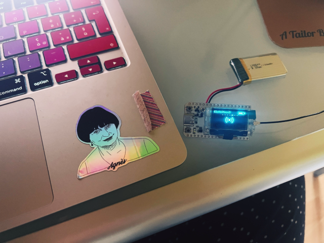 a photo of a laptop with rainbox-color keys and a sticker of Belgian filmmaker Agnès Varda and next to it there is a tiny Heltec v3 unit with a battery plugged in