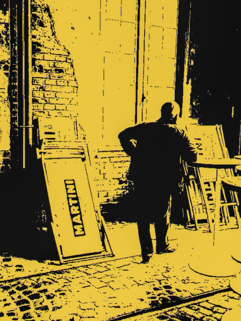 A high-contrast, posterized photograph made with Kodak Chamera film, showing a man standing with his right arm resting on a tall round table outside a former tram depot. Several advertising boards lean against the brick wall behind him, one prominently displaying the word "MARTINI." Tram rails run through the cobblestone street in the foreground. Rendered in bold black and yellow tones, the image has a stark, graphic, almost screen-printed appearance.
