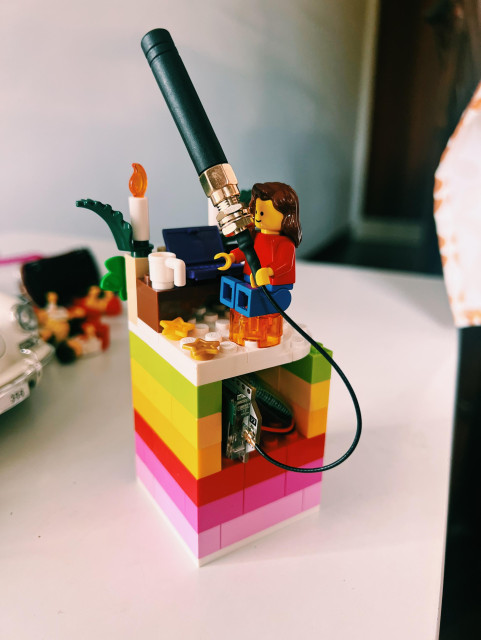 a rainbow-colored LEGO structure with a hole in the middle that houses my Heltec v3 unit and its battery - the antenna cord goes up and it's a minifig on top of the structure - that looks like me - who holds the Heltec antenna in its hand. The minifig is sitting on an orange transparent stool and has a desk with a cup and a purple laptop in front