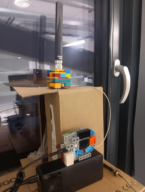Photo with LEGO build fixture for ALPHA LoRA antenna and Heltec V3 Meshcore companion put ontop of a few boxes and powerbank.