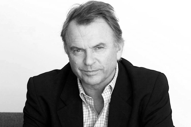 Black-and-white close-up portrait of actor Sam Neill in dark jacket and checked-pattern shirt.