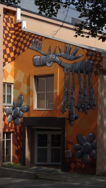 A wall on the corner of a house, painted a bright orange. Around the door and window are two large flowers made of balloons and an animal with six legs but with the body and shape of a moose, painted in a cartoonish style in grey. The creature is grinning and has one eye closed...