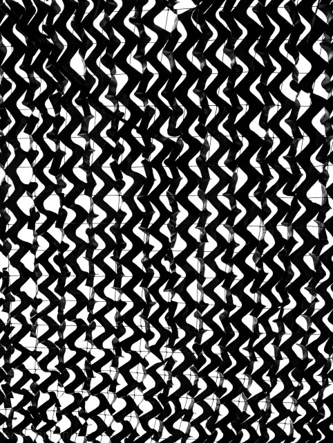 A black-and-white image featuring a dense, repeating abstract pattern that creates a strong optical texture.

Numerous vertical columns of dark, jagged, wave-like or chevron shapes, twist and curve, forming a tightly woven, interlocking mesh.

The silhouette of the camouflage netting is solid black, while the negative spaces (the gaps where light shines through from behind) are a brilliant, stark white.