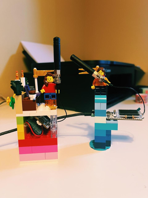 a photo of two LEGO custom builds housing Heltec v3 LORA units. On the left: a rainbow-colored structure with a hole in the middle to accommodate the Heltec and its battery and on top there's a LEGO female minifig with long wavy brown hair holding the antenna. She's sitting at a LEGO desk with a purple laptop and a cup of coffee in front and there are two plants and candles as decorations. Behind there's a gold flag. On the right: a second LEGO structure in various shades of blue housing a second Heltec unit, plugged into power via USB-C. At the top there's a second female LEGO minifig holding the antenna and behind her there's another gold flag. In the background of the photo you can see a black printer