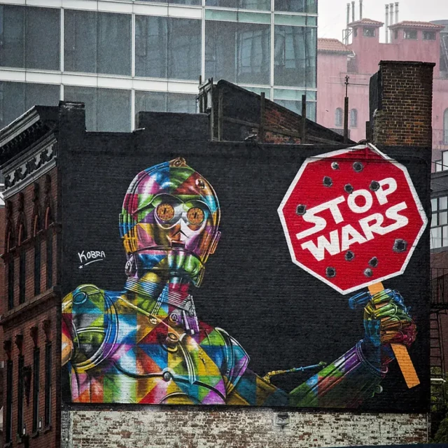 A photo of a huge mural of the Star Wars character C-3PO but in rainbow colours, holding a stop sign riddled with bullet holes.