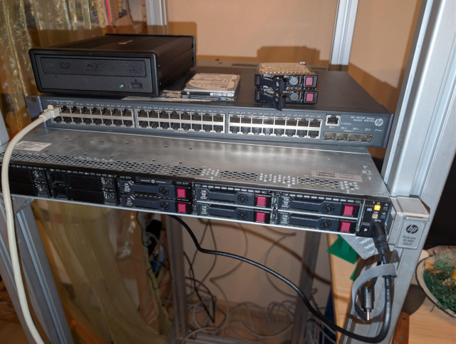 A picture of a part of my homelab. It includes a server (HP Proliant Dl 360 Gen9) in a self-built rack. On top of it, you can find a switch and a Blu-ray player for DVD and Blu-ray ripping. Next to it are the HDDs.