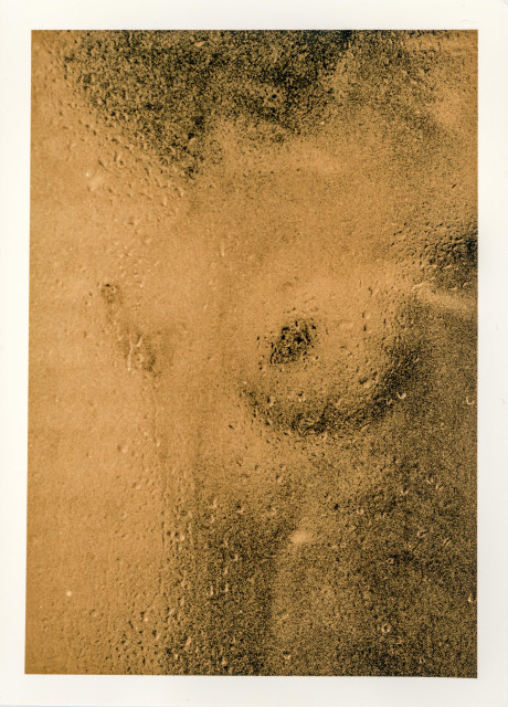This silver gelatin print was produced using the Lith process. It shows a close-up of a portion of a woman’s naked body—her abdomen and breasts. The woman stands behind the glass partition of a shower stall, which is covered with water droplets and steam, blurring her body. The image has a grainy, coarse texture and yellow-brown tones, which are characteristic of lith prints