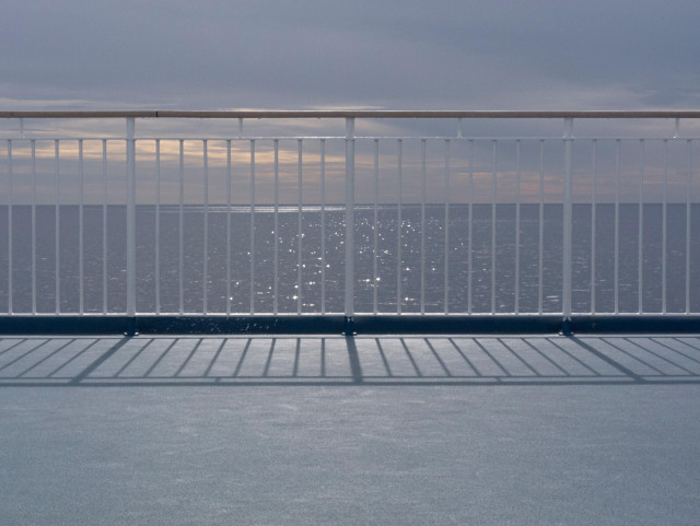 A photo looking over a smooth light blue floor through a white metal railing on a calm sea reflecting the sunlight peeking through a mostly overcast sky.