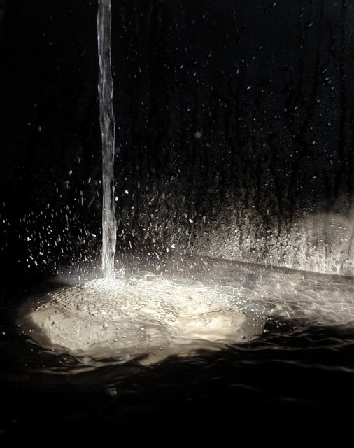 A close-up photo of a water falling from an open tap into a black sink. A spot of sunlight illuminates the white whirl at the bottom.