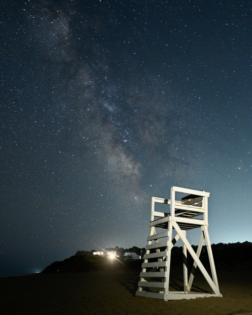 An empty life guard chair stands on the beach. In the background, the lights of a beach house can be seen and, beyond that, the stars of the Milky Way.