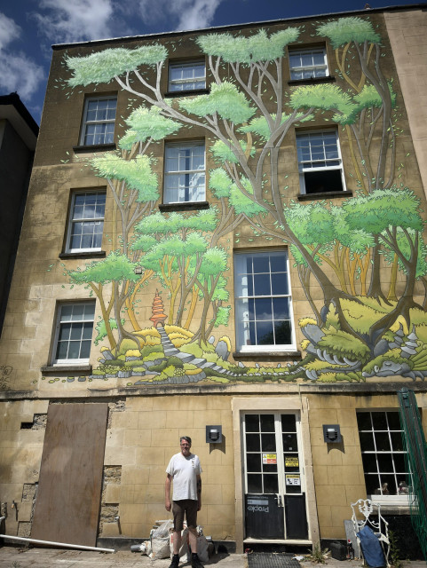 The back wall of a four storey house. The house is 180 years old, paint rendering over stone. On top of this is now a painting of a forest, with bright green trees, with golden tops. At the bottom is a mossy stony ground, with the stones actually a curled, sleeping dragon 