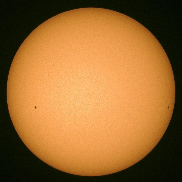 A bright orange sun against a dark background, with two small dark spots visible on its surface indicative of sunspots.