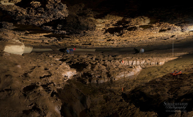 Panoramic style image of a very muddy passage with low ceilings. We can see two figures hunched in a low crawl peering into the channels of water cut through the mud. It is very slippery, difficult not to fall in, yet imperative to not! Lest the water get silted up instantly, or worse, one of the endangered critters in it getting hurt. This cave is "not a fun cave". Not even type 2 fun. Although...it was the day we found this fish!