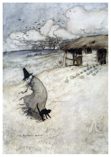 An older woman wearing a pointed witch’s hat stands outside a weathered thatched cottage as a black cat rubs against her legs.