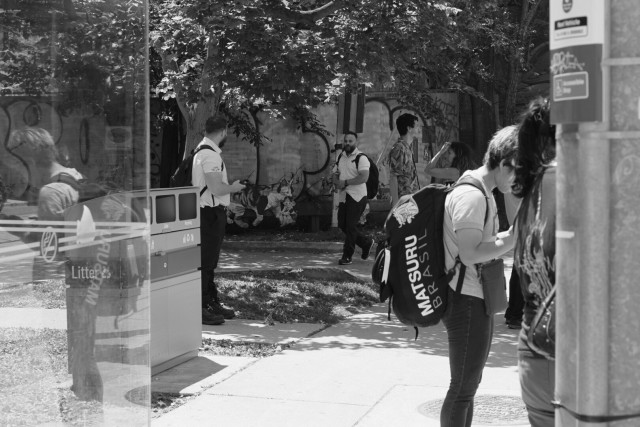 At the streetcar stop, riders wait. A young man wearing a huge backpack looks down at his phone. In the background, two streetcar drivers talk, one passing, one waiting.