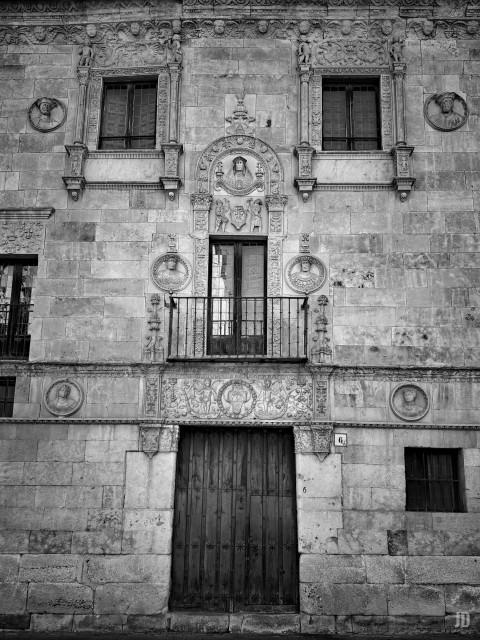 La imagen muestra la fachada de un edificio histórico de piedra, ricamente decorada con relieves y medallones esculpidos que representan rostros humanos. La puerta principal de madera está situada en la parte inferior y sobre ella hay un balcón enrejado con un marco ornamentado. Las ventanas superiores tienen rejas y están rodeadas de molduras talladas con motivos renacentistas. La fachada presenta un estilo clásico y simétrico, destacando su elaboración artística en los detalles arquitectónicos.