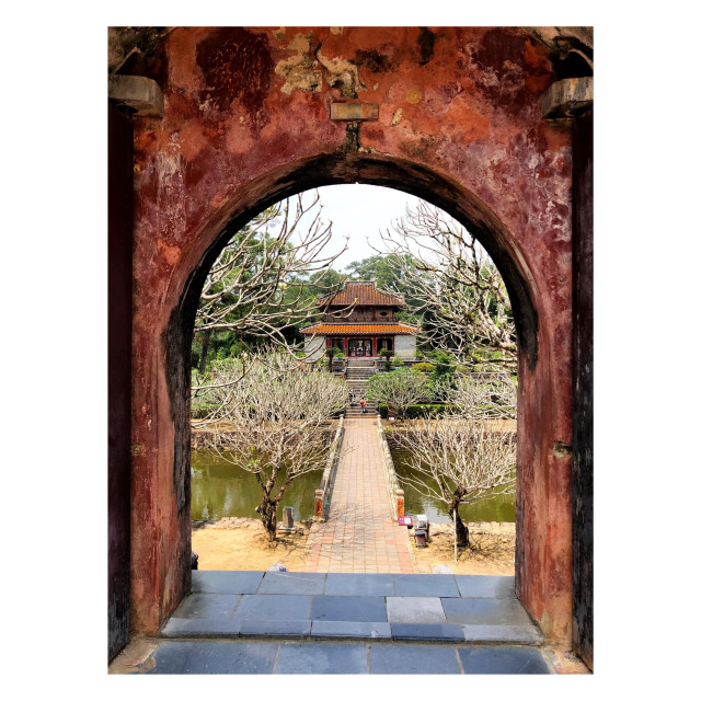 An outdoor view framed by a weathered archway in the foreground with a crumbling, earth-toned surface. A path with large square tiles leads down some steps to a tiled pedestrian bridge that crosses a narrow river. Bare-branched frangipani trees flank the bridge’s near end and push into view from the sides of the arch. On the far side of the river are steps leading up to a two-tiered building with sloped roofs, surrounded by green trees.