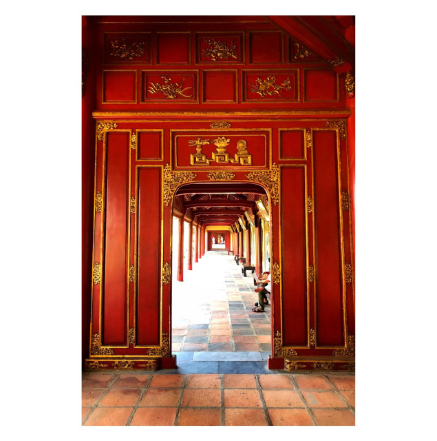 A doorway set into a paneled wooden wall, painted a vivid red with gold borders and ornamentation. A path of brown-orange tiles leads through the door to the outside, where a roof is held up by supporting posts and beams, which appear to the viewer like a series of receding doorways. Bright sunlight from the left side of the path washes out some of the outdoor view.