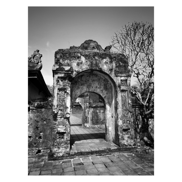 Black and white photo of a weathered outdoor arch with ornamented sides, standing under a cloudless sky. The arch’s crumbling surface bears the marks of time and history, including bullet holes from previous wars. A frangipani tree stands off to its right. A tiled pathway leads through the arch to other arches in the distance.