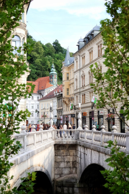 Tromostovje, the Triple Bridge in Ljubljana, Slovenia: a picturesque pedestrian stone bridge comprising three connected spans with balustrades and arches spanning the Ljubljanica River, framed by leafy green trees. Pedestrians walk across towards historic pastel-coloured buildings with ornate Baroque facades and red-tiled roofs. A prominent green-domed clock tower rises above the skyline, while shops and cafes with outdoor seating line the bustling street below. A forested hill, where Ljubljana Castle stands, provides a lush green backdrop under a clear blue sky with scattered white clouds, capturing the charming, lively atmosphere of Ljubljana's old town centre.