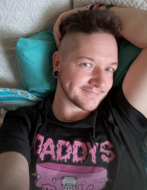 Trans man with dark hair and facial hair. He has blue eyes, and is wearing a black shirt with "Daddy's boy" on it in pink. He reclines against a blue pillow with one arm behind his head, and looks at you with a slightly suggestive smile.