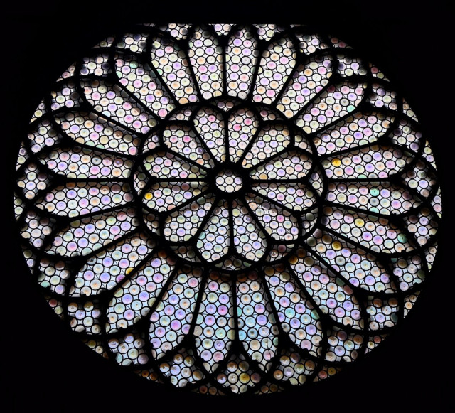 Circular stained glass rose window from Trieste Cathedral, featuring an intricate geometric design composed of numerous small circular panes of iridescent glass in soft pastel hues. Dark lead caming divides the window into radial and concentric patterns, creating a symmetrical, kaleidoscopic effect with subtle colour variations against the black background.