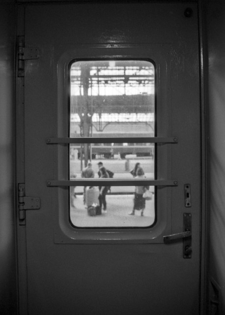 Black-and-white photograph of a train carriage door with a small window framing a blurred railway platform. Several travelers with luggage are visible outside, while the heavy metal door and its horizontal safety bars dominate the composition, creating a quiet sense of separation, departure, and transition.