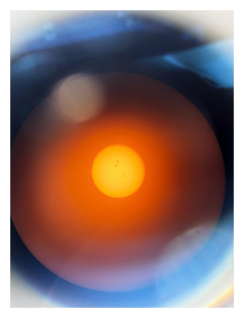 An image of concentric circles. In the middle is the bright orange-yellow disc of the sun, freckled with sunspots. Around it is a red-orange glow, dimming as it reaches the rim of the telescope eyepiece. The rim itself is a thick black circle. Scattered circular afterimages and lens flares appear here and there. The sun’s appearance here strongly evokes the image of the HAL 9000 computer from the film “2001.”