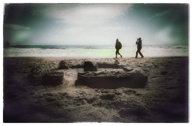 A picture taken on Rodeo Beach of a sand fort: a circular ring wall surrounding a central structure, with an open entryway land-side. In the distance, closer to the shore, two silhouetted people have noticed but do not yet realize they growing danger. Crabs do not take prisoners.
