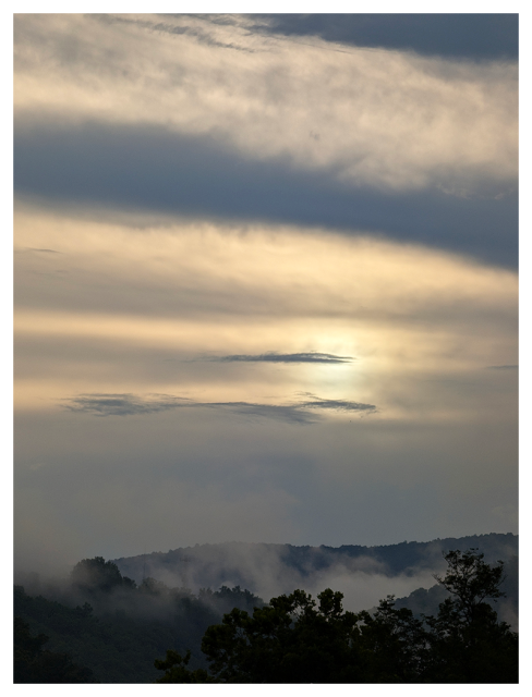	
clouds and mist hang over rolling hills at sunset, with the sun partially obscured behind light clouds. the sky transitions from pale yellow near the sun to soft gray-blue higher up, while trees and fog line the dark foreground.