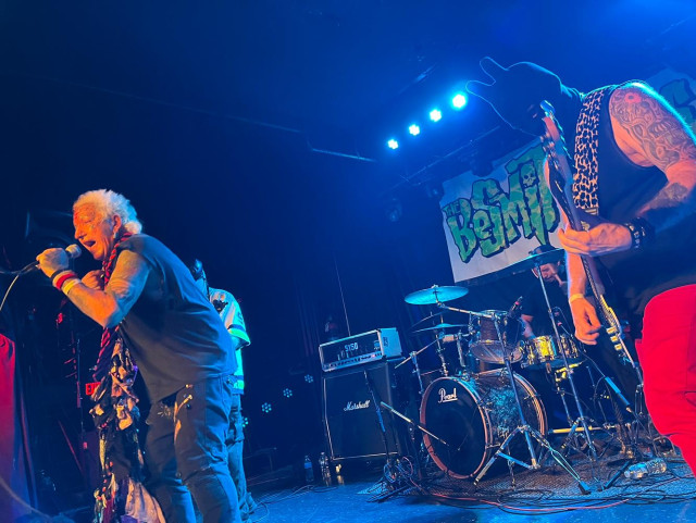 Besmirchers band performing live on a small stage. The vocalist is wearing cut-up clothes and has lots of tattoos and piercings. The bass player is wearing a bunny-shaped ski mask. The other two members are slightly cloaked and there is a giant banner with their logo behind them.