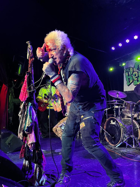 Lenny singing. His face and hand are bloody and he’s grimacing with passion. He has white, spikey hair, lots of tattoos and piercings, and is wearing cut-up clothes and jeans with zippers all over them. His mic stand is dripping with colorful cloth.