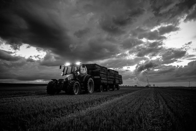 Ein Traktor zieht einen Anhänger mit Heuballen über ein abgeerntetes Feld. Er hat die beiden Lampen oben am Traktor an, der Himmel ist stark bewölkt. Im Hintergrund ist ein Kraftwerk zu erkennen. Das Foto ist in schwarz-weiß. 