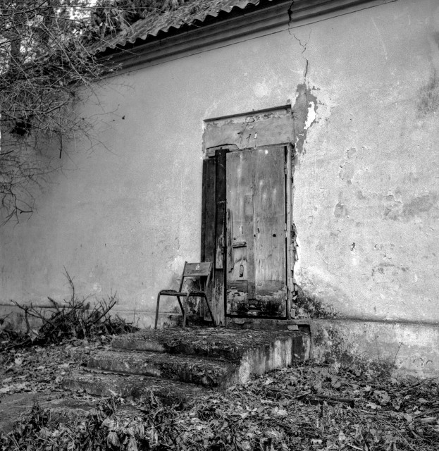 A black-and-white photograph of a weathered building with a cracked plaster wall and a worn wooden door at the top of a few crumbling concrete steps. A single old chair rests beside the entrance, while dry leaves, bare branches, and overgrown vegetation surround the neglected structure, creating a quiet, abandoned atmosphere.