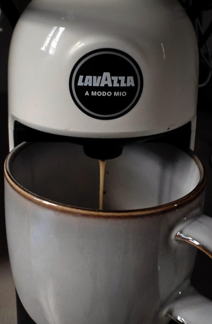 A close-up photo showing a small Lavazza coffee machine with a stream of espresso falling into a white ceramic mug with a brown rim. 