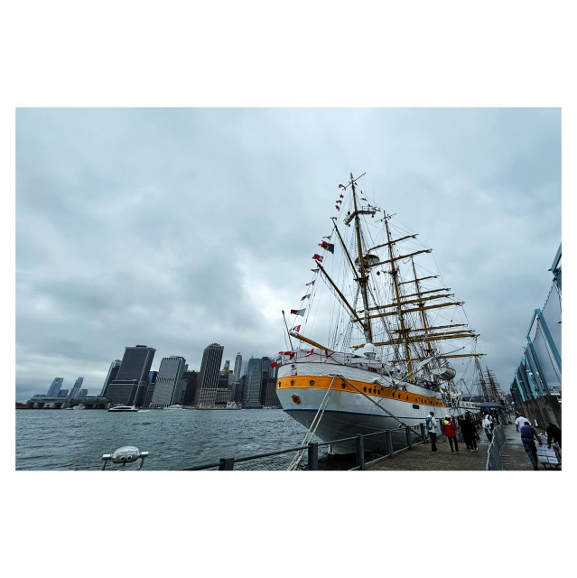 A large three-masted barque with its sails furled floats next to a pier with people milling about outside and on the deck. Its hull is white with a broad horizontal yellow stripe. In the image it’s off-center to the right, and behind it is the East River with the skyline of lower Manhattan. The sky is overcast with gray clouds.