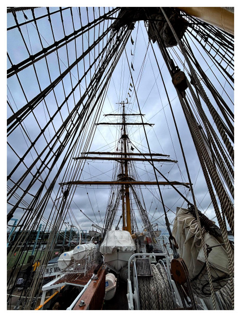 A vertical wide-angle view onboard the deck of the tall ship Mircea, looking up towards the rear mast. The vertical pole and horizontal spars — as well as the diagonal ropes of the rigging radiating out from the central mast that’s just above and behind camera — create an image of striking geometric lines and angles. On the deck is a jumble of coiled ropes, furled sails, and covered lifeboats.