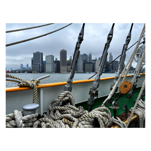 A view of lower Manhattan from the deck of the tall ship Mircea. The deck has a yellow top railing with white and green sections below. A jumble of slack ropes lies on deck while other ropes extend upwards past the frame. Three long metal cylinders lean diagonally rightward and appear to connect some rope lines to the deck. Past them we can see the East River with Manhattan’s buildings on the far side, some of their tops disappearing into fog in an overcast sky.