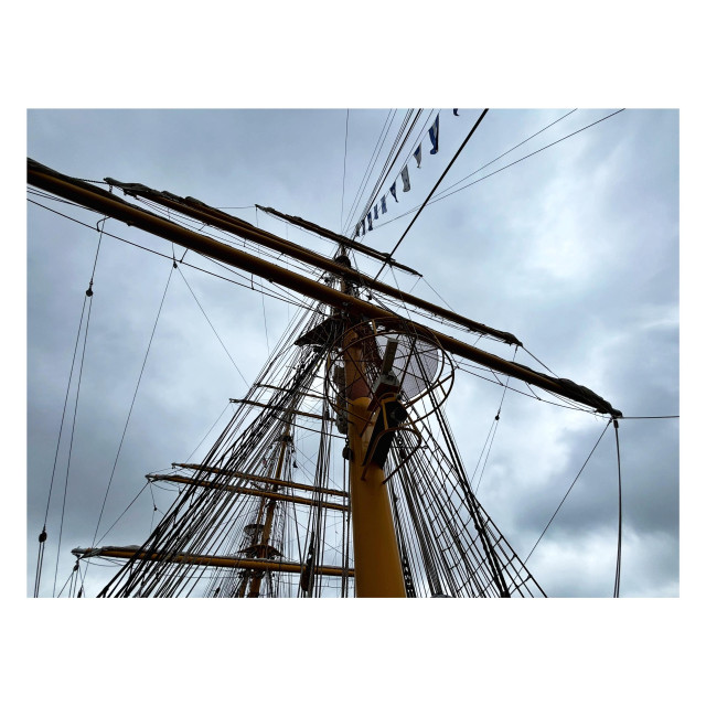 An angled view looking up at one of the masts of the tall ship Mircea. Three horizontal spars cross the vertical pole, though they’re angled slightly diagonally because of the orientation of the camera. Rope lines crisscross and connect with the spars at various points, and a mesh of rigging descends from the mast’s midpoint to the deck. One line of rope carries a series of small banners. The sky is overcast with gray clouds.