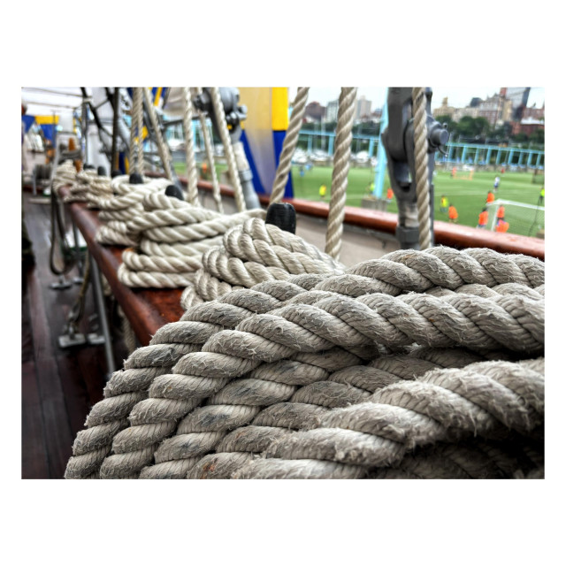 Closeup of several coils of rope lined up along the wooden railing of the tall ship Mircea. Beyond are some blue-and-yellow banners. Blurred in the distance are soccer players on a green field, which is adjacent to the pier where the ship is docked.