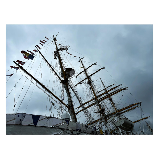 Another angled view looking up at the tall ship Mircea, this one including all three of its masts as well as a lifeboat hanging off its side. The ship is backlit by vague sunlight in an overcast sky. Small banners attached to one of the rope lines flutter in the breeze.