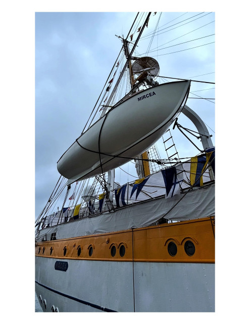 View looking up at a lifeboat hanging off the side of the tall ship Mircea. We see the underside of the boat, its hull painted white with “MIRCEA” in blue letters near its prow. Below is the larger ship’s hull with a broad yellow stripe running across its portholes and a series of blue-and-white banners along its top. Behind the lifeboat, one of the ship’s masts rises into a cloudy sky.