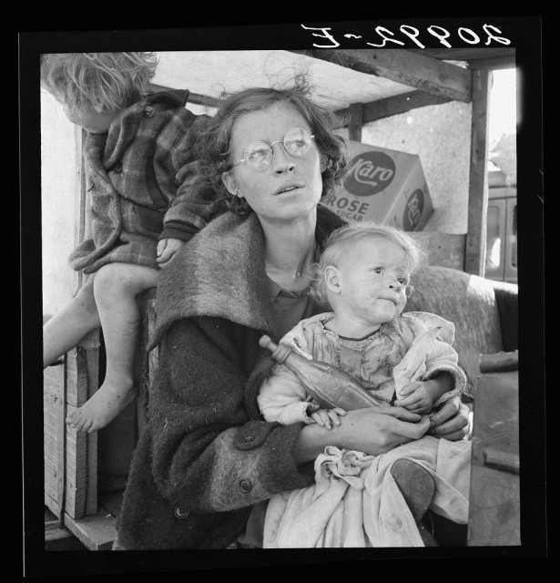 The image depicts a black and white photograph featuring an older woman with glasses, wearing a heavy coat. She is holding two children; one in her arms appears to be looking directly at the camera while the other child is seated on her lap. The environment suggests they are outside near some sort of wooden structure or booth where items like "Karo" syrup and "Rose Cigar" paper can be seen, indicating a possible setting during an economic hardship period.

The woman's expression appears to convey exhaustion or contemplation as she gazes off into the distance. The children appear malnourished and dressed in worn-out clothing, reflecting poverty. This photograph is likely from historical documentation of impoverished families during difficult times, possibly around the Great Depression era given the attire and conditions depicted.