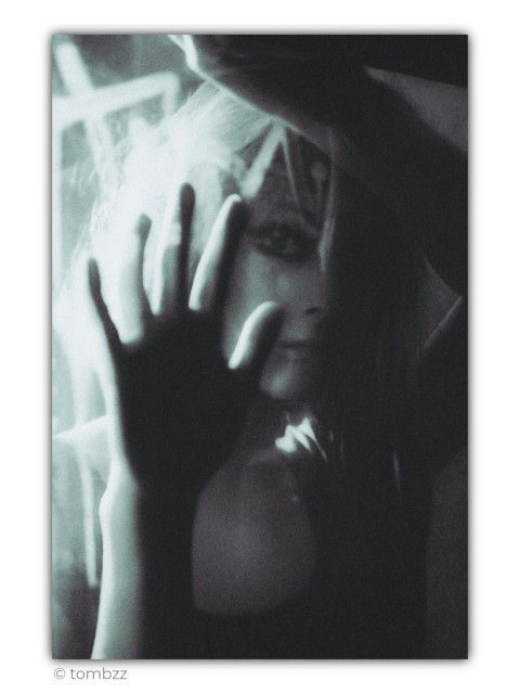Black-and-white analog portrait in a noir aesthetic. A young woman stands behind a pane of glass, resting both hands against it. One hand, closest to the camera, dominates the foreground—dark, slightly out of focus, and partially obscuring her face, creating a strong visual barrier between the subject and the viewer. Her other hand is raised above her head, framing the composition from the top. Between them, part of her face remains visible, with one eye looking directly into the lens. The gaze is calm yet intense, carrying a quiet sense of unease. Her bold eye makeup gives the eyes even greater presence, drawing attention despite the soft tonal transitions.
The lighting is deliberately overexposed. Bright highlights around her hair and along the edges of the glass almost dissolve into white, while the right side of the frame falls into deep shadow. The image doesn't strive for technical perfection. Its softness, subtle blur, strong contrast, and analog grain create a dreamlike, cinematic atmosphere. This is a portrait where mood takes precedence over precision—a photograph suspended somewhere between reality and memory, inviting the viewer to find their own interpretation.