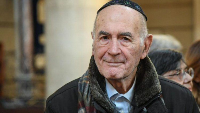 Elderly man wearing a kippah and scarf, attending an indoor event.
