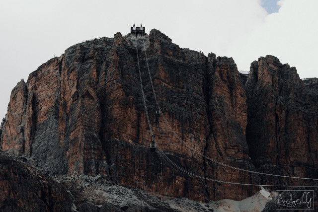 Rock face (Sass Pordoi) with a long steep cable car chain and a small platform at the top under a cloudy sky