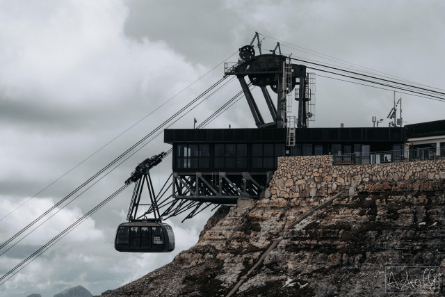 Cable car gondola suspended on steel cables next to a mountain station on rocky terrain under cloudy sky