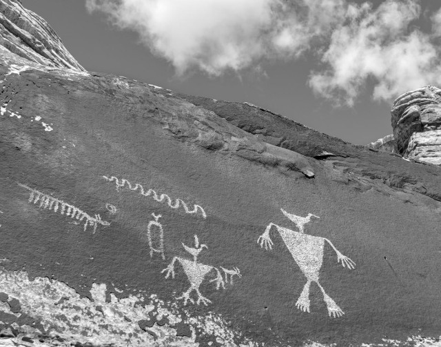 A black and white landscape photo of rock imagery on a very dark desert vanish background. There are seven figures pecked into the flat rock face. The figures are light colored against the natural dark vanish of the rock. The main figure on the right looks like a person with flipper feet and a duck for a head. To the lower left is a smaller image similar to the large one on the right except this one also has a small four legged animal near its right shoulder. Further to the left is an oval with short legs and another duck looking head. Above that is a horizontal squiggle line. And on the far left is possibly the image of a centipede. (Note: I only describe these images in terms that my modern eye sees them. To the person, man, woman, or child, who drew them they very likely had entirely different representations and meanings. These images are likely 800 - 1,000 years old and none of us are of the culture that made them so what they are and what they mean is now lost to time. Also, the term "rock imagery" is preferred by the local tribes to "rock art" since not even they know the meaning or purpose of their ancestor's anicent petroglyphs and pictographs. Referring to the images as "art" is therefore considered disrespectful.)