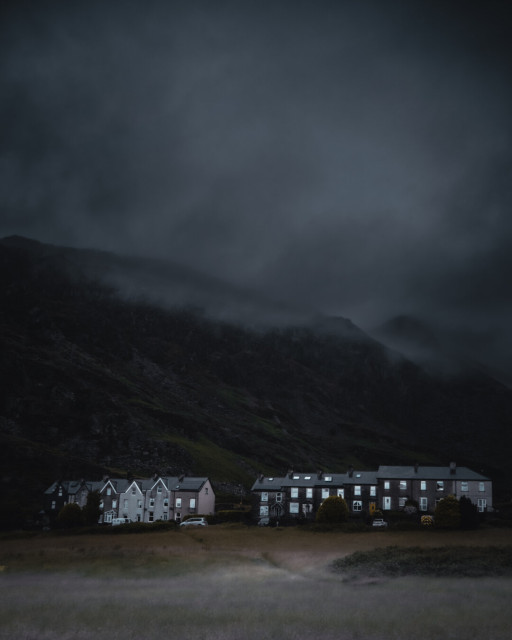 A low-light photo of a remote mountain village. Two small sets of terraced houses with glowing windows stand in a dim valley, in the shadow of a dark, towering mountain shrouded in thick low-hanging fog and storm clouds.