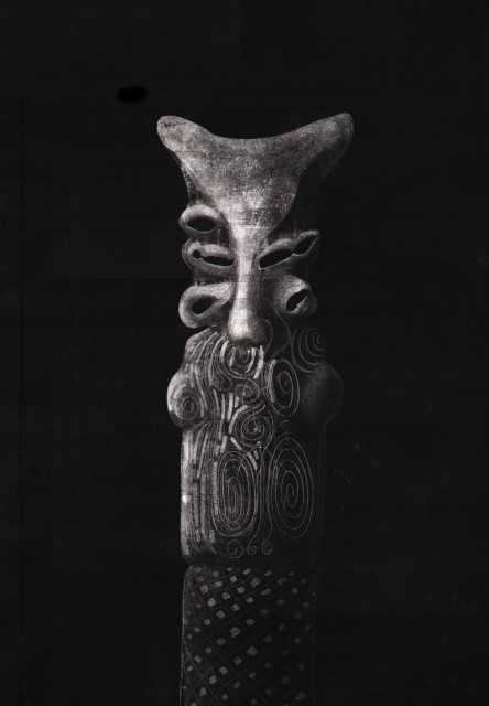 A black and white scratchboard drawing of a vertical anthropomorphic totem or fetish figure, characterized by intricate textures. The top of the figure is anvil-shaped, positioned above a mask-like face with concave eye sockets and a central nose/mouth structure. The mid-section features a dense pattern of intertwined spirals and figure-eight motifs, while the base displays a geometric grid of textured diamonds and hexagons. The entire piece is rendered against a deep, dark black background.
